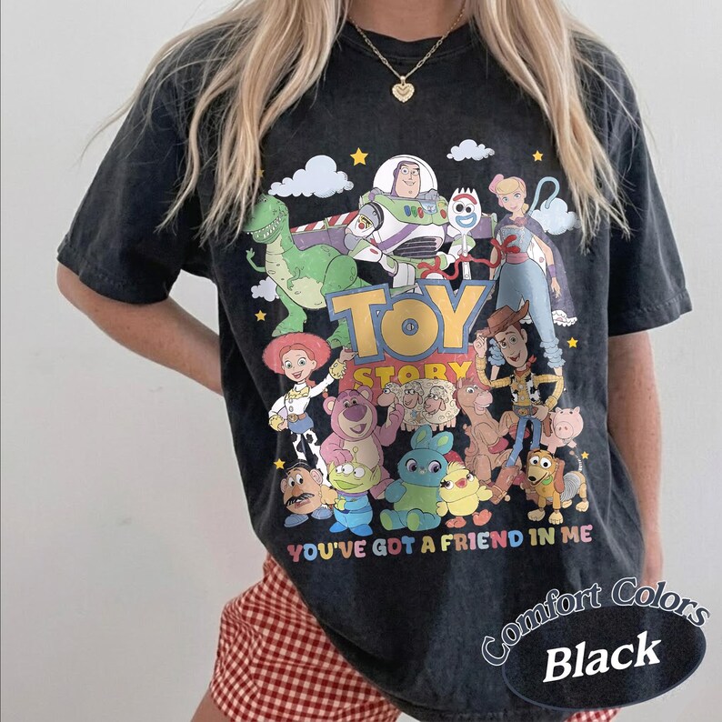 May include: A black short-sleeve t-shirt featuring a colorful graphic print of Toy Story characters including Buzz Lightyear, Woody, Jessie, and Rex. The text Toy Story and You've Got A Friend In Me are printed in the center with small stars and clouds.