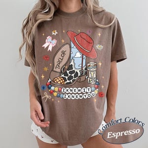 May include: A brown short-sleeve t-shirt featuring a graphic print of a cowboy boot with a cow print pattern, a red hat, building blocks with the letters T, S, and a cloud design, a bow, and beads. The text reads Taylor, 13, I KNEW IT, I KNEW YOU.
