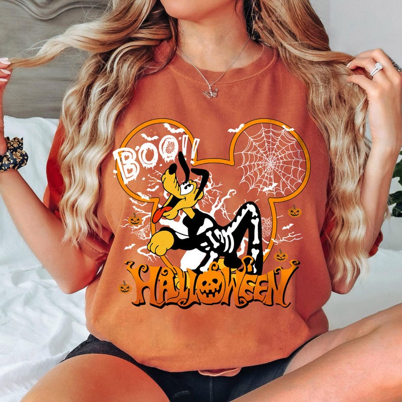 May include: Orange t-shirt with a Halloween-themed graphic featuring Pluto in a skeleton costume, a spider web, and the words "BOO!" and "HALLOWEEN". The design is set within a Mickey Mouse head outline.