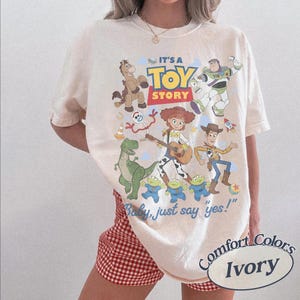 May include: An ivory short-sleeve t-shirt featuring a colorful graphic print of Toy Story characters including Woody, Buzz Lightyear, Jessie, Rex, Bullseye, Forky, and the aliens. The text reads IT'S A TOY STORY and Baby, just say yes!.