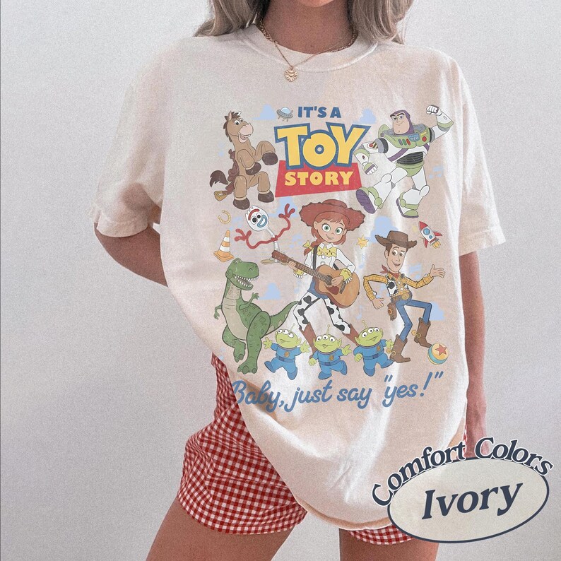 May include: An ivory short-sleeve t-shirt featuring a colorful graphic print of Toy Story characters including Woody, Buzz Lightyear, Jessie, Rex, Bullseye, Forky, and the aliens. The text reads IT'S A TOY STORY and Baby, just say yes!.