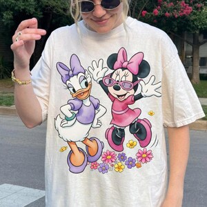 May include: Cream-colored t-shirt featuring Daisy Duck and Minnie Mouse. Daisy Duck is in a purple outfit, and Minnie Mouse is in a pink dress and sunglasses. Both characters are surrounded by colorful flowers.