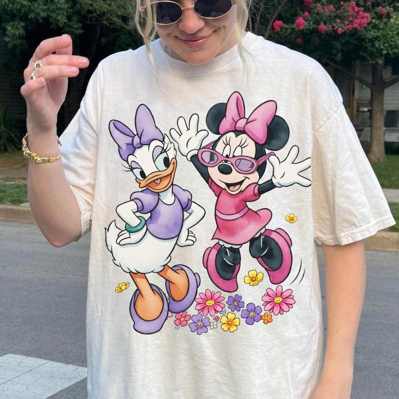 May include: Cream-colored t-shirt featuring Daisy Duck and Minnie Mouse. Daisy Duck is in a purple outfit, and Minnie Mouse is in a pink dress and sunglasses. Both characters are surrounded by colorful flowers.