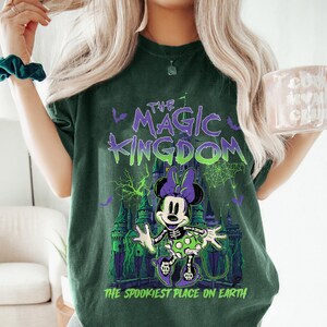 May include: Dark green t-shirt featuring a Halloween-themed graphic of Minnie Mouse in a skeleton costume in front of a castle. The text "THE MAGIC KINGDOM" and "THE SPOOKIEST PLACE ON EARTH" are printed on the shirt.