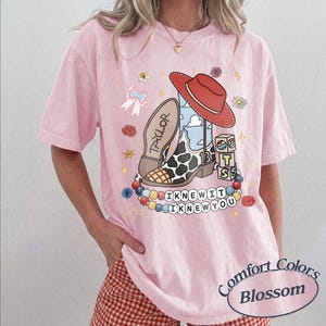 May include: A light pink t-shirt featuring a graphic print of a cowboy boot with a cow print pattern, a red cowboy hat, alphabet blocks, and a string of colourful beads. The text "TAYLOR", "13", and "I KNEW IT I KNEW YOU" are printed on the shirt.