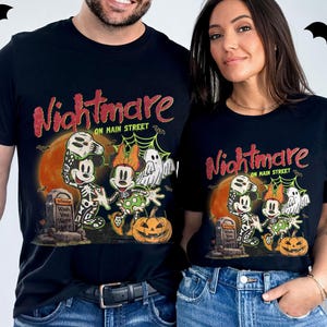 May include: Black t-shirts with a Halloween-themed graphic. The design features Mickey and Minnie Mouse as skeletons, a ghost, and a jack-o'-lantern, with the words "Nightmare on Main Street". Bats are in the background.
