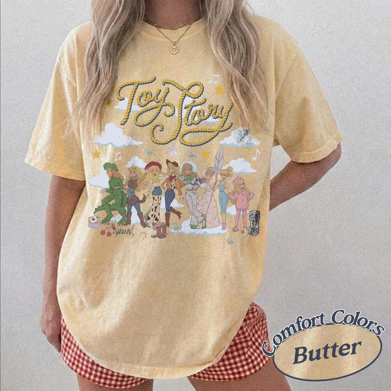 May include: A pale yellow short-sleeve t-shirt featuring the text Toy Story in a rope-style font. Below the text is a graphic illustration of various animated characters standing among clouds, stars, and musical notes. The shirt has a crew neckline.