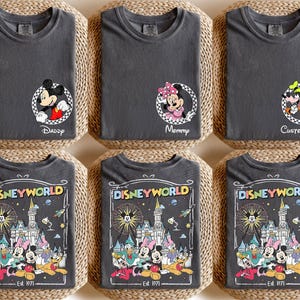 Op de afbeelding: Zes antracietgrijze t-shirts met Disney-ontwerpen. Drie shirts hebben Mickey, Minnie en Goofy in cirkelvormige frames met tekst. De andere drie shirts tonen een kleurrijke Disney World-afbeelding met personages en tekst.