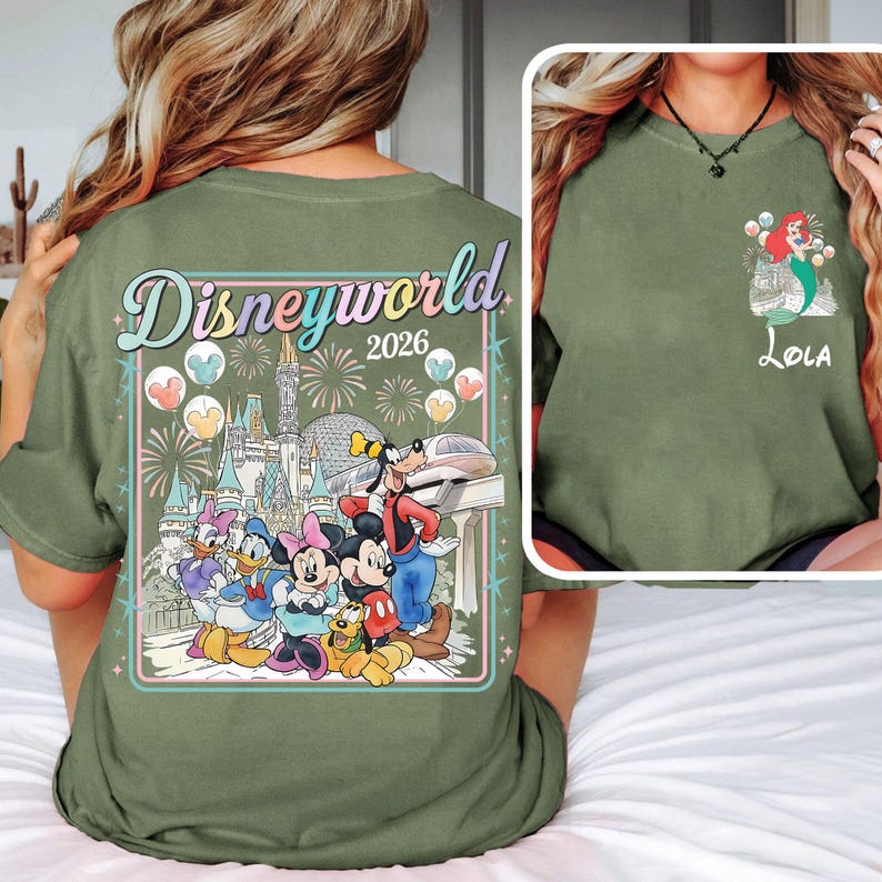 Pu&ograve; includere: T-shirt verde oliva con un design Disneyworld 2026. Il design include personaggi Disney, fuochi d'artificio e un castello. &Egrave; presente anche una piccola immagine di Ariel con il nome "Lola".