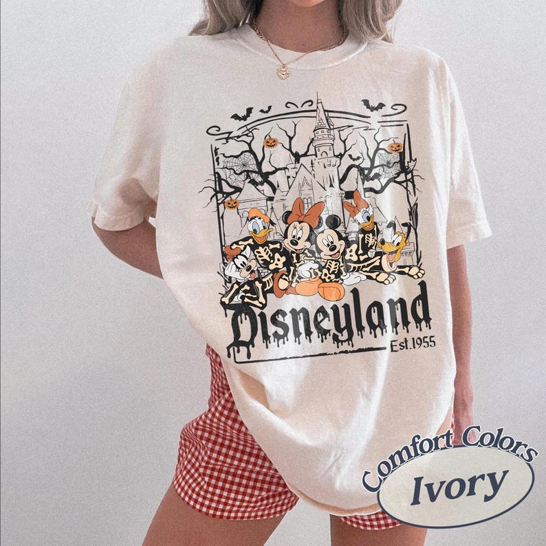 May include: Ivory-colored t-shirt featuring a Disneyland graphic with cartoon characters in skeleton form. The design includes a haunted castle, bats, and pumpkins. The text "Disneyland Est. 1955" is printed below the characters.