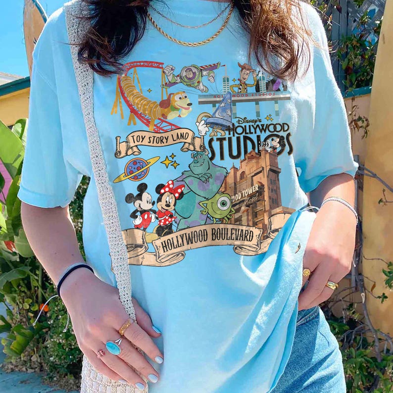 May include: Light blue t-shirt featuring a colorful graphic of Toy Story Land and Hollywood Studios. The design includes cartoon characters, a roller coaster, and the text "Hollywood Boulevard".