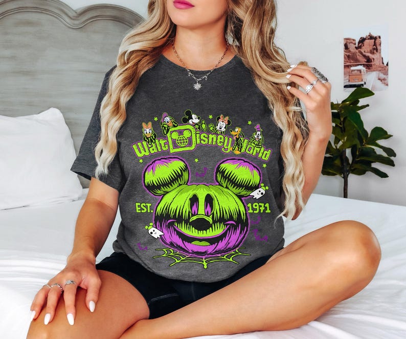 May include: Dark gray t-shirt featuring a Halloween-themed design with a green and purple Mickey Mouse pumpkin graphic. The text "Walt Disney World" and "EST. 1971" are also visible. The shirt has a crew neck and short sleeves.