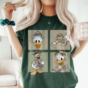 May include: Dark green t-shirt with a four-panel design of Donald Duck in safari clothing. Each panel shows a different pose of Donald Duck wearing a safari hat and outfit. The background panels have a striped and textured design.