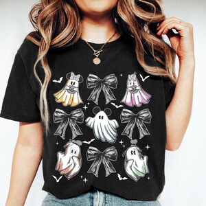 May include: Black t-shirt featuring a Halloween-themed design. The design includes cartoon ghosts with bows, bats, and stars. The ghosts are in various colors, including yellow, pink, and white. The bows are black.