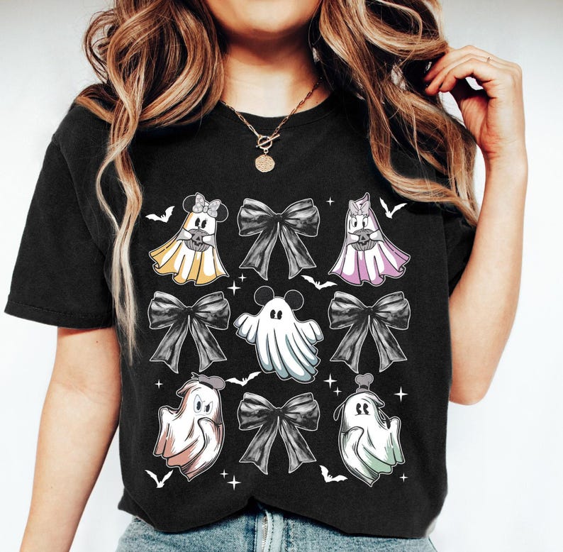 May include: Black t-shirt featuring a Halloween-themed design. The design includes cartoon ghosts with bows, bats, and stars. The ghosts are in various colors, including yellow, pink, and white. The bows are black.