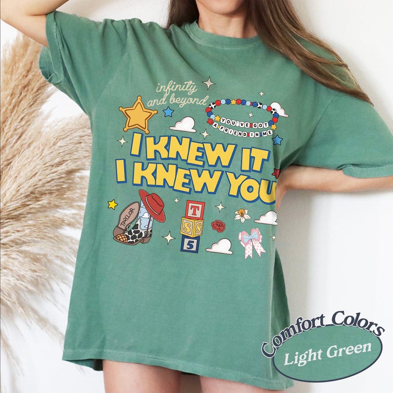 May include: A sage green t-shirt featuring a graphic print with the text "infinity and beyond", "I KNEW IT I KNEW YOU", and "YOU'VE GOT A FRIEND IN ME". The design includes a yellow star, a beaded bracelet, a cowboy boot, alphabet blocks, and a bow.