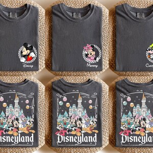 May include: Six charcoal gray t-shirts with Disney character designs. Some shirts feature Mickey and Minnie Mouse, and others display a Disneyland graphic with the text "Disneyland 1955". The shirts are folded on woven mats.