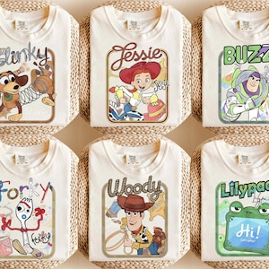 May include: Six off-white t-shirts featuring graphic prints of characters including Slinky, Jessie, Buzz Lightyear, Forky, Woody, and a frog labelled Lilypad. Each shirt displays a rectangular frame with character names and illustrations.