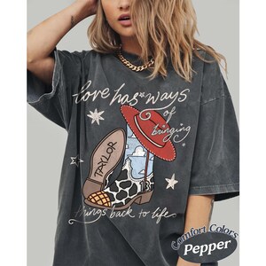 May include: A charcoal gray t-shirt featuring a graphic of a cowboy boot with a cow print pattern, a blue sky print, and the number 13, topped with a red hat. The text reads Love has ways of bringing things back to life and Taylor.