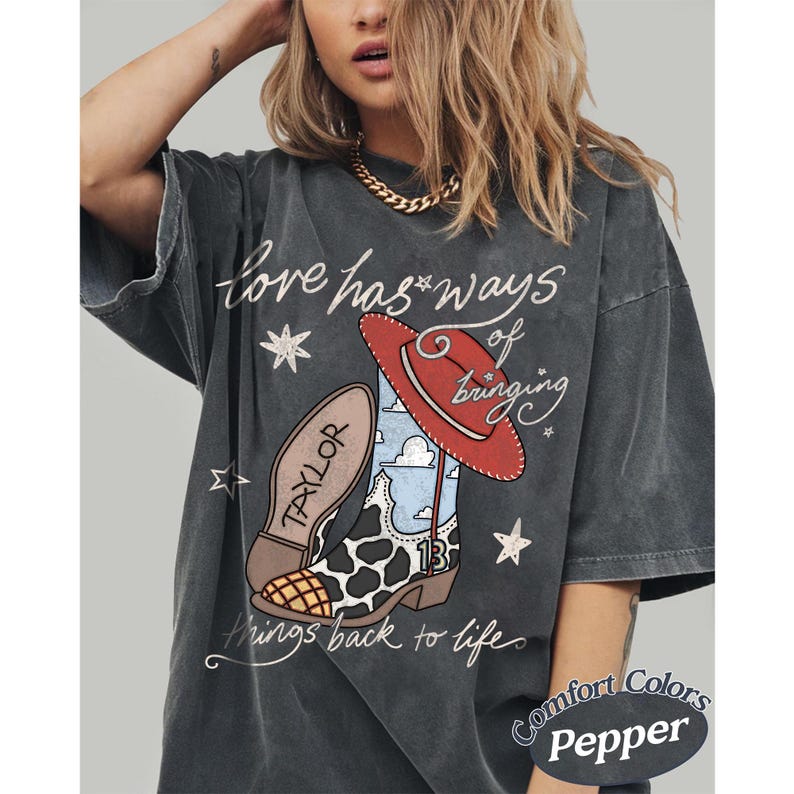 May include: A charcoal gray t-shirt featuring a graphic of a cowboy boot with a cow print pattern, a blue sky print, and the number 13, topped with a red hat. The text reads Love has ways of bringing things back to life and Taylor.