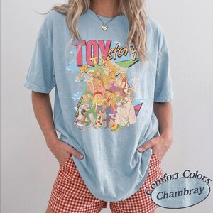 May include: A light blue t-shirt featuring a graphic print with the text TOY story. The design displays a colorful illustration of various animated characters in a group, including figures in western and space-themed outfits, set against a geometric background.