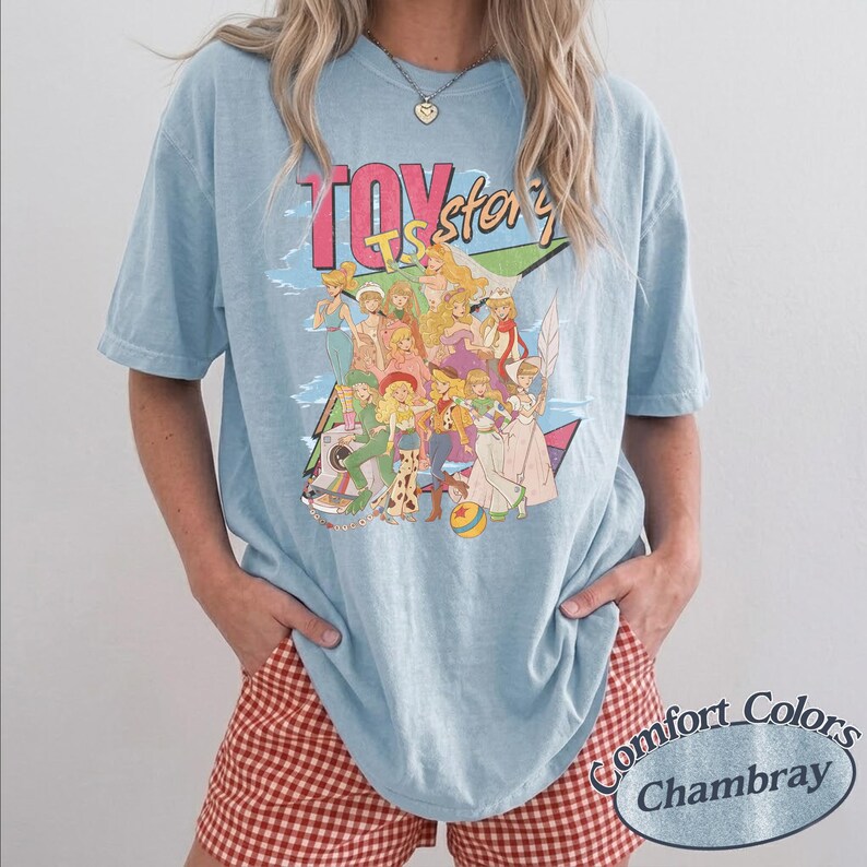 May include: A light blue t-shirt featuring a graphic print with the text TOY story. The design displays a colorful illustration of various animated characters in a group, including figures in western and space-themed outfits, set against a geometric background.