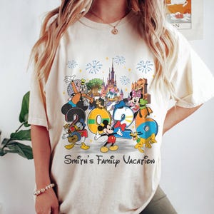 Może przedstawiać: Kremowy t-shirt z kolorową grafiką postaci Disneya i numerem "2026". Projekt zawiera Myszkę Miki, Kaczora Donalda, Goofy'ego i Minnie, z fajerwerkami i zamkiem w tle. Tekst "Smith's Family Vacation" jest wydrukowany poniżej.