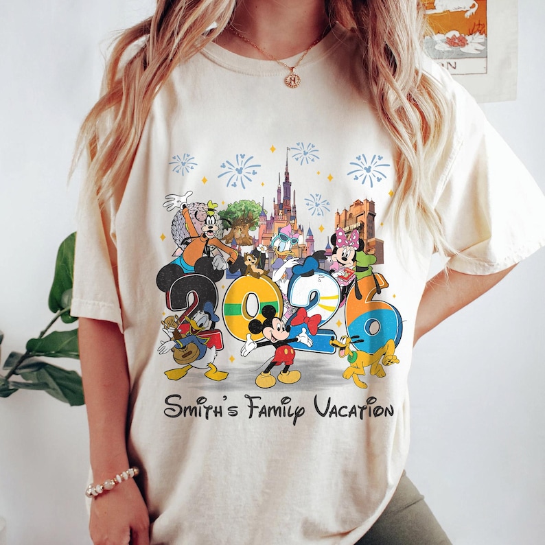 Może przedstawiać: Kremowy t-shirt z kolorową grafiką postaci Disneya i numerem "2026". Projekt zawiera Myszkę Miki, Kaczora Donalda, Goofy'ego i Minnie, z fajerwerkami i zamkiem w tle. Tekst "Smith's Family Vacation" jest wydrukowany poniżej.