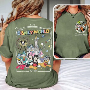 Op de afbeelding: Olijfgroen T-shirt met een Disney World-ontwerp op de achterkant, met Mickey Mouse, Minnie Mouse en vrienden. De voorkant heeft een Goofy-afbeelding. Tekst bevat "Disneyworld" en "Est. 1971".