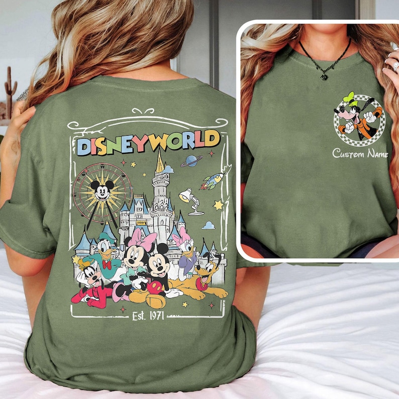 Op de afbeelding: Olijfgroen T-shirt met een Disney World-ontwerp op de achterkant, met Mickey Mouse, Minnie Mouse en vrienden. De voorkant heeft een Goofy-afbeelding. Tekst bevat "Disneyworld" en "Est. 1971".