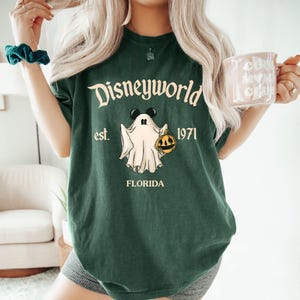 Op de afbeelding: Groen T-shirt met een witte geest die een pompoen vasthoudt en de tekst "Disneyworld est. 1971 Florida".