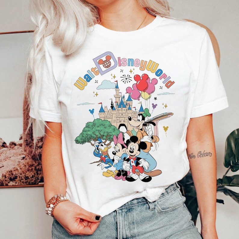 May include: White t-shirt with a colorful graphic of Mickey Mouse, Minnie Mouse, Donald Duck, and Goofy in front of Cinderella's Castle. The text "Walt Disney World" is written in a rainbow arc above the characters.