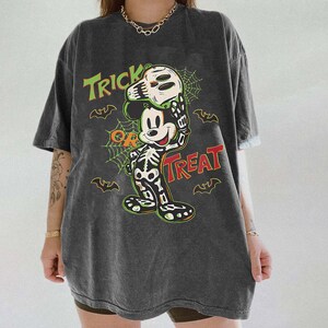 May include: Dark grey t-shirt with a Halloween-themed Mickey Mouse design. The graphic shows Mickey in a skeleton costume holding a skull, with the words "Trick or Treat" in green and red. Bats and spiderwebs complete the design.