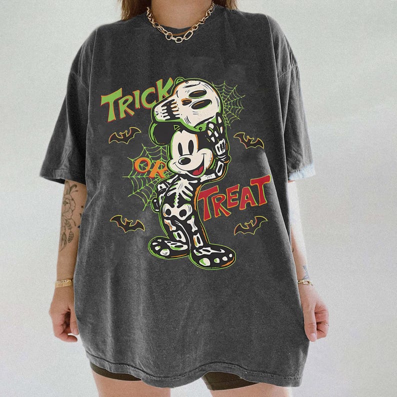 May include: Dark grey t-shirt with a Halloween-themed Mickey Mouse design. The graphic shows Mickey in a skeleton costume holding a skull, with the words "Trick or Treat" in green and red. Bats and spiderwebs complete the design.