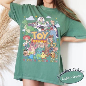 May include: A light green short-sleeve t-shirt featuring a colorful graphic print of Toy Story characters including Woody, Buzz Lightyear, and Jessie. The text Toy Story and You've Got a Friend in Me are printed in the center of the shirt design.