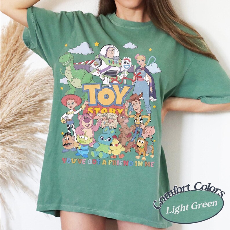 May include: A light green short-sleeve t-shirt featuring a colorful graphic print of Toy Story characters including Woody, Buzz Lightyear, and Jessie. The text Toy Story and You've Got a Friend in Me are printed in the center of the shirt design.