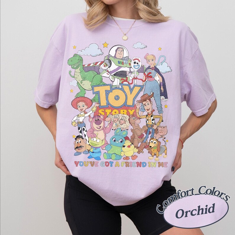 May include: A light purple short-sleeve t-shirt featuring a colorful graphic print of Toy Story characters including Woody, Buzz Lightyear, and Rex. The text Toy Story and You've Got A Friend In Me are printed on the front. The shirt has a crew neckline.