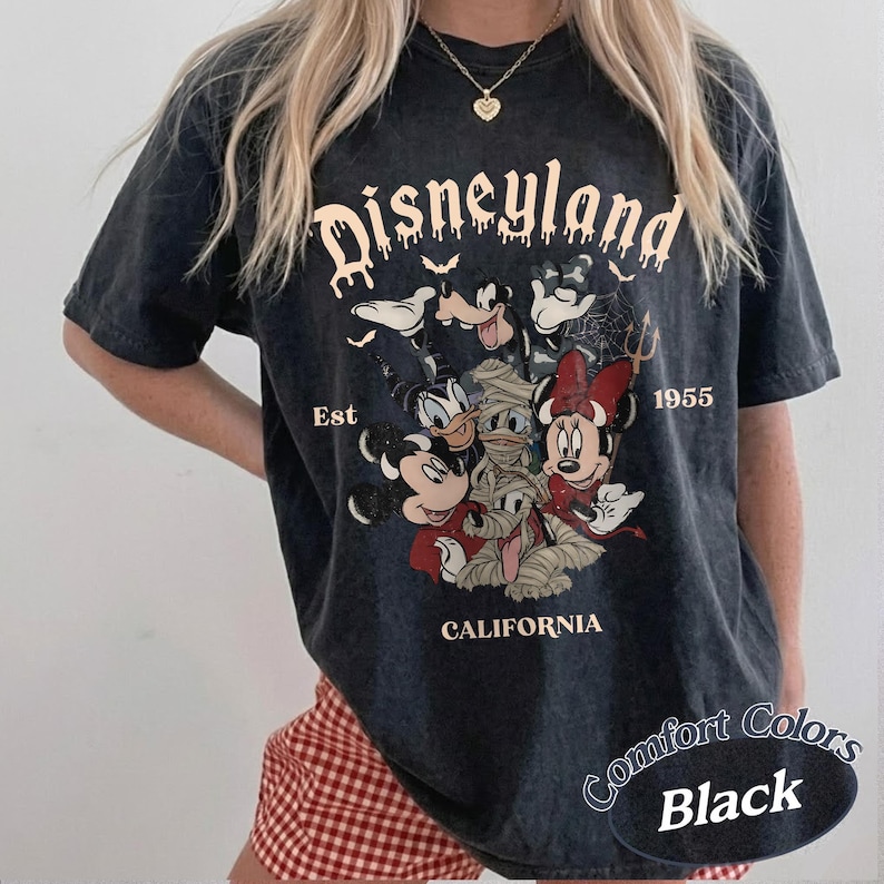 May include: A black mineral-washed t-shirt featuring a graphic print of Disney characters including Mickey, Minnie, Donald, and Goofy in Halloween costumes. The text reads Disneyland Est 1955 California. The shirt has short sleeves and a crew neckline.