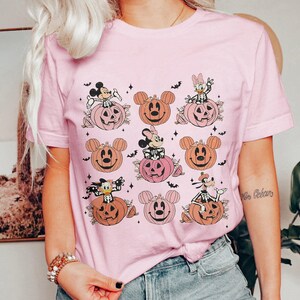 May include: Pink t-shirt featuring a Halloween-themed design. The graphic includes cartoon characters like Mickey Mouse, Minnie Mouse, Donald Duck, and Goofy, each depicted as skeletons sitting on pumpkins. Bats and stars complete the design.