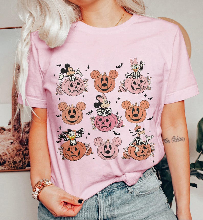 May include: Pink t-shirt featuring a Halloween-themed design. The graphic includes cartoon characters like Mickey Mouse, Minnie Mouse, Donald Duck, and Goofy, each depicted as skeletons sitting on pumpkins. Bats and stars complete the design.