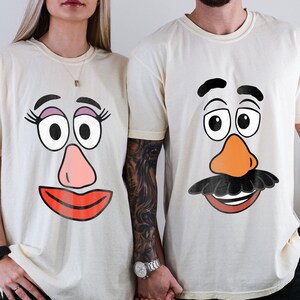 May include: Ivory t-shirts with cartoon faces. One shirt shows a female face with pink lips, a pink nose, and large eyes. The other shirt has a male face with an orange nose and a black moustache.