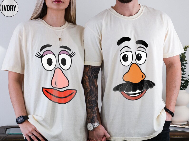 May include: Ivory t-shirts with cartoon faces. One shirt shows a female face with pink lips, a pink nose, and large eyes. The other shirt has a male face with an orange nose and a black moustache.