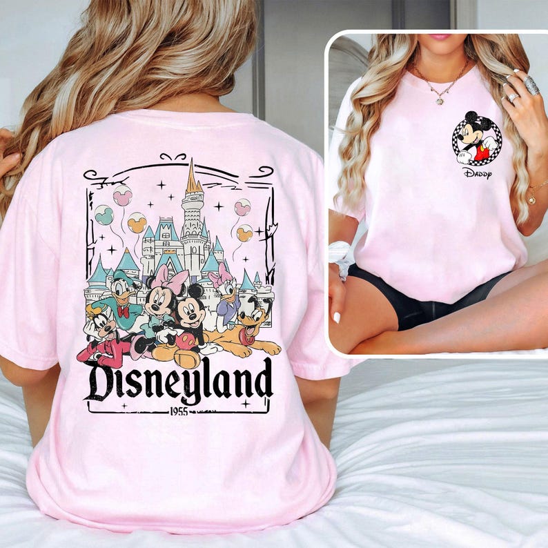 May include: Light pink t-shirt featuring a Disneyland graphic on the back with Mickey Mouse, Minnie Mouse, Donald Duck, Daisy Duck, Goofy, and Pluto. The front of the shirt has a Mickey Mouse graphic. The text "Disneyland" is displayed on the back.