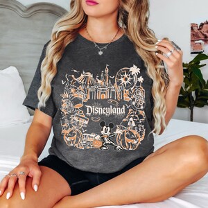 May include: Dark gray t-shirt featuring a Disneyland design with orange and white graphics. The design includes a castle, Ferris wheel, pumpkins, and Mickey Mouse. The word "Disneyland" is prominently displayed.