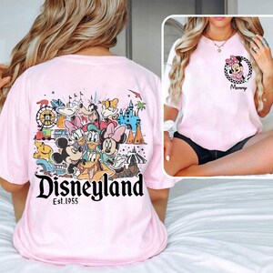 May include: Pale pink t-shirt with a Disneyland graphic featuring Mickey Mouse, Minnie Mouse, Donald Duck, and other Disney characters. The shirt also has the text "Disneyland Est. 1955". A smaller image of Minnie Mouse is on the front.