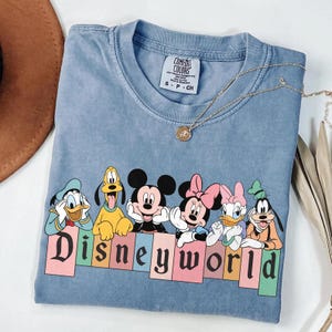 Pode incluir: Uma camiseta azul claro com um gr&aacute;fico vintage de Mickey Mouse, Minnie Mouse, Donald Duck, Pateta e Pluto. Os personagens est&atilde;o em p&eacute; em frente a uma placa colorida "Disney World".