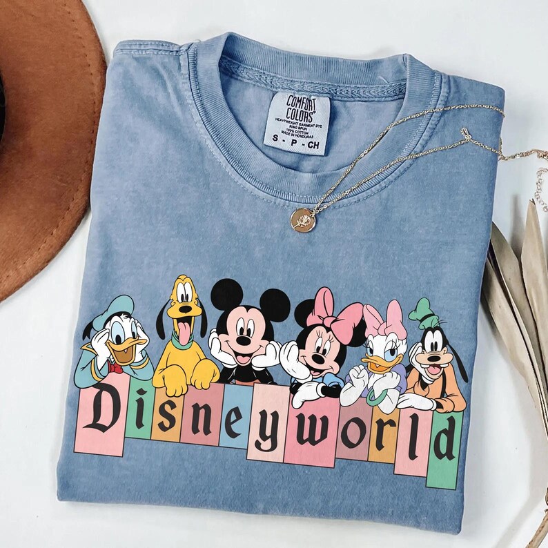 Pode incluir: Uma camiseta azul claro com um gr&aacute;fico vintage de Mickey Mouse, Minnie Mouse, Donald Duck, Pateta e Pluto. Os personagens est&atilde;o em p&eacute; em frente a uma placa colorida "Disney World".