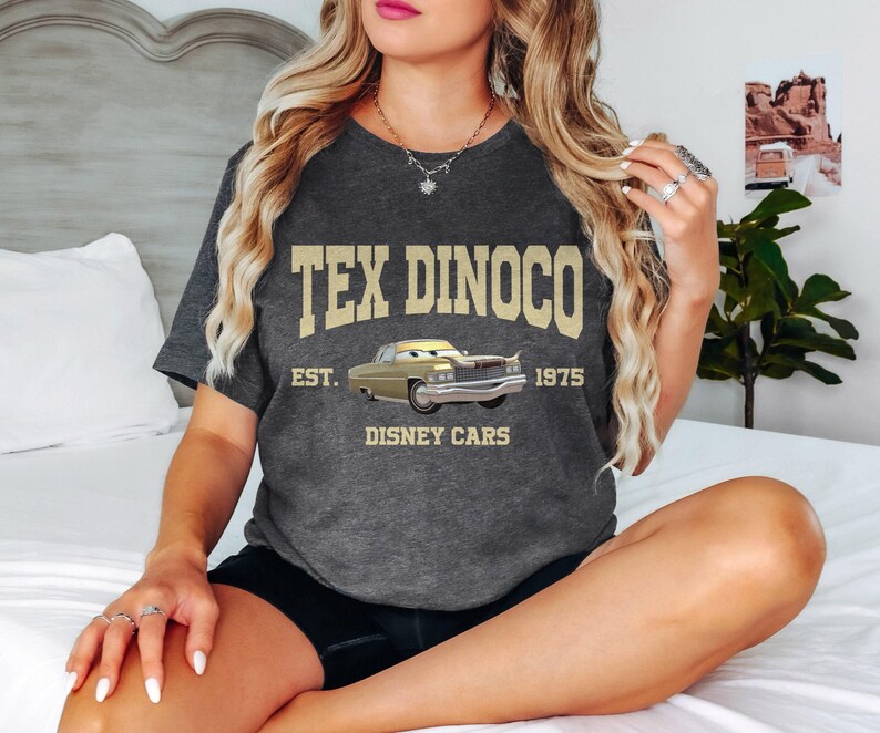 Puede incluir: Una camiseta gris oscuro con el texto "Tex Dinoco Est. 1975 Disney Cars" en letras doradas. Un coche de dibujos animados aparece en el centro de la camiseta.