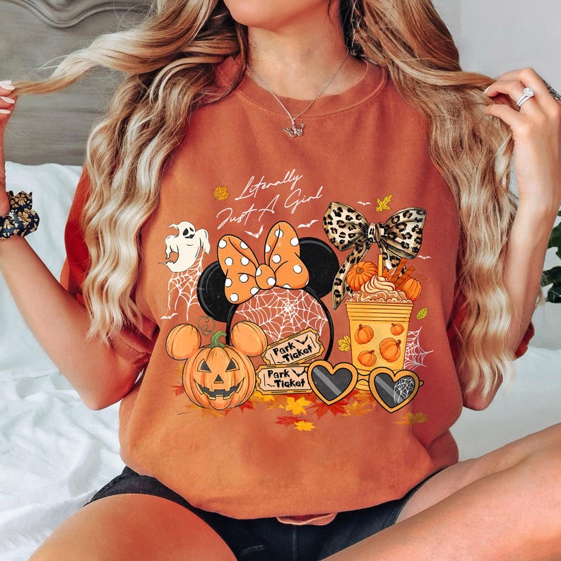 May include: Burnt orange t-shirt with a Halloween-themed graphic. The design includes a Minnie Mouse head with a polka dot bow, a jack-o'-lantern, a ghost, and the text "Literally Just A Girl".