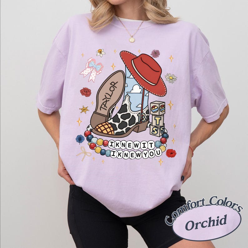 May include: A light purple t-shirt featuring a graphic of a cow-print cowboy boot with a red hat, a bow, blocks with the letters T, S, and a star, and a beaded necklace with the text I KNEW IT I KNEW YOU. The boot has the name TAYLOR printed on it.